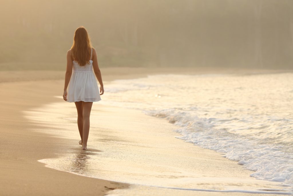 Set bagfra af en kvinde, der går på sandet ved stranden ved solnedgang